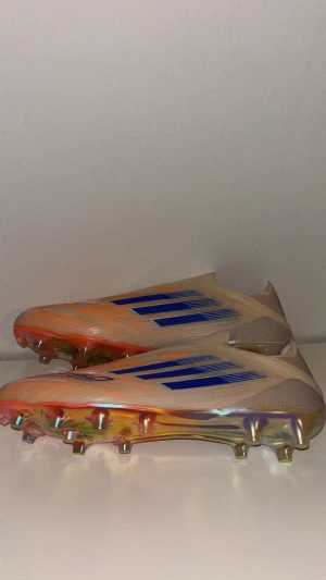 Adidas F50 SPARKFUSION | CORAL BLAZE  - Säljer ett par Adidas F50 fotbollsskor i beige med blå och orange detaljer. Skorna har en strömlinjeformad design utan snörning, med platt sula och mönstrad ovandel. Yttersulan har en skimrande finish och dobbar för gräsplan. Perfekt för dig som vill sticka ut på planen.