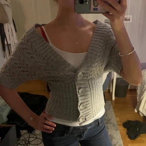 Grå stickad kofta med kort ärm - Superfin grå stickad kofta/bolero med korta ärmar och bred ribbad kant. Koftan har stora grå knappar framtill och ett snyggt mönster i stickningen. Sitter tajt i midjan och har en djup v-ringning. Perfekt att styla över ett linne eller t-shirt.