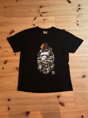 Svart BAPE t-shirt med camo print - Svart t-shirt från BAPE med ikoniskt ape-motiv på bröstet i grönt, brunt och beige camouflage. Klassisk kortärmad modell i mjuk bomull. BAPE-logga på ärmen och färgstark etikett i nacken. Perfekt för dig som gillar streetwear och unika prints.
