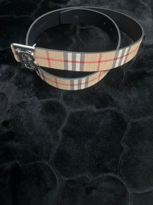 Burberry bälte med silverdetalj - Snyggt bälte från Burberry med klassiskt rutigt mönster i beige, rött, svart och grått. Bältet har en stilren silverfärgad spänne med Burberrys ikoniska TB-logga. Perfekt accessoar för att lyfta din outfit med en lyxig touch.