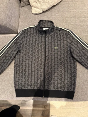 Svart Lacoste tröja L - Snygg svart bomberjacka från Lacoste med geometriskt mönster och vita ränder längs ärmarna. Jackan har dragkedja framtill, ribbade muddar och klassisk Lacoste-logga på bröstet. Regular fit och sportig vibe, perfekt för dig som gillar streetstyle. Kvitto finns!
