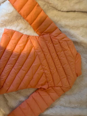 Orange/rosa pufferjacka från Haust Collection - Snygg orange/rosa pufferjacka från Haust Collection med broderad logga på insidan och patch på ärmen. Jackan har två dragkedjeförsedda fickor, hög krage med knappdetalj och quiltad design. Perfekt för dig som vill sticka ut med färg i vinter.