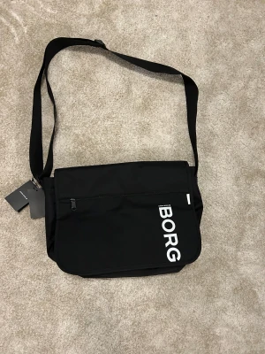 Svart axelväska från Björn Borg - Stilren svart axelväska från Björn Borg med justerbar rem, stort huvudfack och praktisk dragkedja framtill. Väskan har aldrig blivit använd. Den har tydlig logga i vitt och är tillverkad i slitstark polyester. Perfekt för att bära med sig laptop, böcker eller andra prylar. Nypris 350kr.