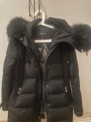 Hollies jacka - Säljer en svart pufferjacka från Hollies med stor huva och fluffig päls. Jackan har guldiga dragkedjor, knappar och detaljer samt logga på ärmen. Inte för kort utan den täcker om man vill sitta ned. 