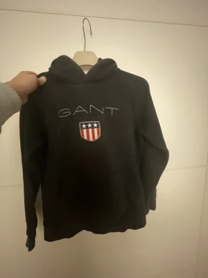 Svart hoodie från GANT med tryck - Svart hoodie från GANT med broderad logga och amerikanskt inspirerat emblem på bröstet. Tröjan har huva, magficka och långa ärmar. Perfekt för dig som gillar klassisk streetwear med snygga detaljer.