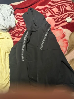 Svart hoodie Icono Couture - Svart hoodie från Icono Couture med huva och snörning. Snygga detaljer med logotypband längs axlarna och ett litet emblem på bröstet. Tröjan har en stor magficka och är tillverkad i mjuk bomull. Perfekt för chill eller streetwear.