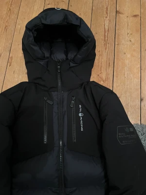 Äkta Sail Racing dunjacka med huva i svart - Säljer en svart dunjacka från Sail Racing, modell Antarctica Expedit eller partoll down jacket. Puffad design och riktigt varm, perfekt för kalla dagar. Den har 5 fickor med två vid midjan och två på bröstet som det får plats mycket I samt så finns det en ficka inne i jackan vid bröst höjd ungefär.  Ny pris på jackan är 3600kr, jag har haft den i typ 3 månader. Den har ett litet hål men man ser ingen skillnad. Jag har tröttnat på den. PRIS KAN DISKUTERAS            Skriv för mer info ✅