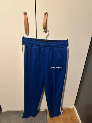 Palm Angels byxor - Snygga blå mjukisbyxor från Palm Angels med vita ränder längs sidorna och logotyp broderad på framsidan. Byxorna har elastisk midja, raka ben och sidofickor. Perfekt för en chill och sportig look.