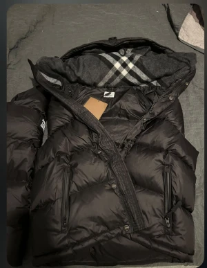 Svart pufferjacka från Burberry - Säljer en svart pufferjacka från Burberry med klassiskt rutigt mönster i huvan. Jackan har dragkedja, knappar och två sidofickor med dragkedja. Materialet är polyester och jackan har en quiltad design som ger en schysst volym. Perfekt för kalla dagar. Priset går att diskutera✌️🫡