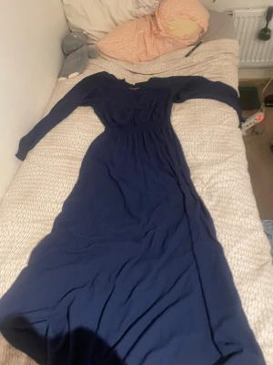 Mörkblå långklänning från H&M - Långklänning i mörkblått från H&M med lång ärm, v-ringning med gulddetalj och smockad midja. Klänningen har en enkel och stilren design, perfekt för dig som gillar klassiska plagg med en twist. Materialet känns mjukt och följsamt.