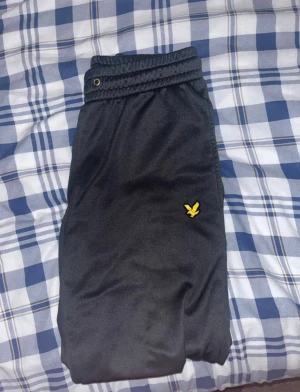 Svarta mjukisbyxor från Lyle & Scott - Svarta mjukisbyxor från Lyle & Scott med elastisk midja och det klassiska gula logobroderiet på benet. Perfekta för chill dagar eller när du vill ha en avslappnad stil. Mjukt material och bekväm passform.