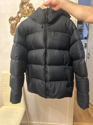 Svart pufferjacka från Peak Performance - Säljer en svart pufferjacka från Peak Performance med huva och två sidofickor med dragkedja. Jackan har en quiltad design och är tillverkad i syntetmaterial, perfekt för kalla dagar. Stängs med dragkedja framtill och har en clean, oversized look.