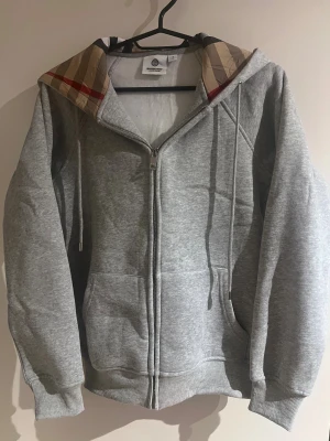 Burberry zip up  - Snygg grå hoodie från Burberry med klassiskt rutigt mönster i huvan och silverfärgad dragkedja med Burberry-logga. Tröjan har långa ärmar, snörning vid huvan och två fickor framtill. Perfekt för dig som gillar stilrena och exklusiva plagg.
