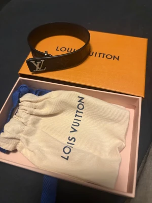 Louis Vuitton brunt armband med LV-logo - Snyggt brunt armband från Louis Vuitton med klassisk LV-logo i silverfärgad metall. Armbandet har diskret monogrammönster och levereras i originalask och tygpåse. Perfekt accessoar för dig som gillar lyxiga detaljer. Armbandet är äkta och nypris ligger runt 3899kr. 10/10 skick knappt använts. Kan skickas/mötas upp redan idag.