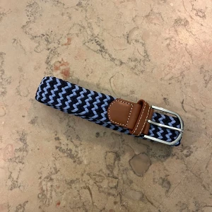 LjusBlåt och marinblått randigt flätat bälte - Snyggt flätat bälte med ett randigt mönster i mörkblått och ljusblått. Spännet är i silverfärgad metall och ändarna är förstärkta med brunt skinn. Perfekt för att ge outfiten lite extra edge och passar till jeans eller chinos.