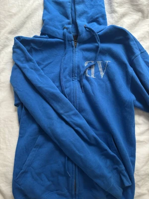 Blå hoodie med strass från Bvaldi  - Blå hoodie från Bvaldi med dragkedja och huva. Snygga strassdetaljer med bokstäver på bröstet och ryggen. Tröjan har snörning vid huvan och är tillverkad i mjuk bomullsmix. Nypris 1300