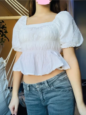 Vit blus med puffärmar och volang - Superfin vit blus från Pernilla Wahlgren med puffiga korta ärmar, fyrkantig ringning och smockad midja. Blusen är croppad med volang nedtill och har en liten guldfärgad knappdetalj. Har aldrig använt