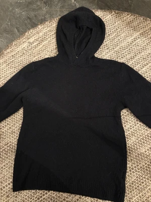 Svart stickad hoodie med huva - Svart stickad hoodie med huva och långa ärmar. Tröjan har en enkel och clean look utan detaljer eller mönster, perfekt för dig som gillar basic och stilrent. Materialet är mjukt och skönt stickat, passar dig som vill ha en chill och bekväm vibe.