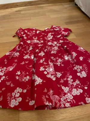 Röd blommig klänning från Lindex Kids - Supersöt röd klänning från Lindex Kids med vita blommor, fjärilar, kaniner och hjortar som mönster. Klänningen har korta ärmar, knappar bak och en utställd kjol. Perfekt för dig som gillar djur och naturinspirerade prints.