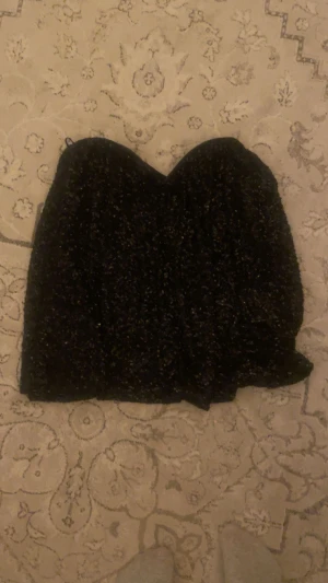 Svart glittrig bandeau tubtopp - Säljer en svart glittrig bandeau tubtopp med hjärtformad urringning. Toppen är axelbandslös och har en loose passform som faller snyggt över kroppen. Perfekt för dig som vill sticka ut med lite extra glitter. Fråga gärna om fler bilder eller mått!