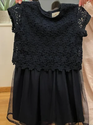 Svart spetsklänning Holly & Whyte - Supersöt svart kortklänning från Holly & Whyte med blommig spets upptill och luftig kjol i flera lager. Klänningen har korta ärmar och rund halsringning. Perfekt för dig som gillar en klassisk men ändå trendig look.