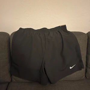 Oanvända Svarta Nike Challenger träningsshorts - Nike Challenger shorts i svart med klassisk vit swoosh-logga på benet. De har elastisk midja och är tillverkade i lätt polyester som andas. Passformen är normal och längden är 7 tum, perfekt för träning eller sport. Snygg och enkel design som funkar till gymmet eller löprundan.