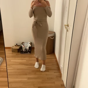 Beige ribbad offshoulder klänning - Säljer en beige, ribbad långklänning med offshoulder och lång ärm. Klänningen har en snygg knytning i midjan som framhäver figuren och ger en trendig look. Materialet är stickat och stretchigt, vilket gör den både bekväm och stilren.