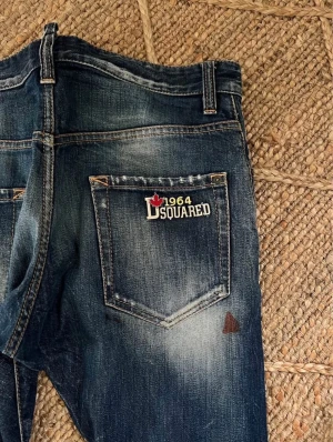 Dsquared2 blå slitna jeans - Snygga blå jeans från Dsquared2 med coolt sliten look, patch med logga och lönnlöv på bakfickan. Jeansen har raka ben, tydliga slitningar och hål framtill samt ljusa tvättade partier. Perfekt för dig som gillar street och edgy stil.
