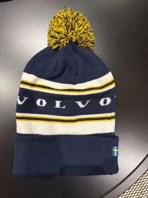 Volvo mössa med tofs och ränder - Snygg mörkblå stickad mössa från Volvo med gul och vit randigt mönster, stor tofs på toppen och svensk flagga på kanten. Perfekt för kalla dagar och visar din Volvo-stolthet på ett stilrent sätt.