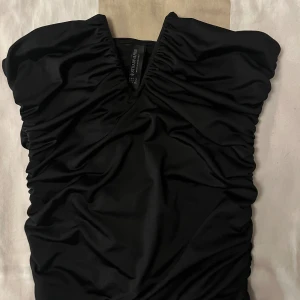 Svart topp från Stradivarius - Supersnygg svart kort klänning från Stradivarius med v-ringning och draperade detaljer längs sidorna. Klänningen är axelbandslös och har en tight passform som framhäver figuren. Perfekt för dig som gillar en stilren och trendig look.