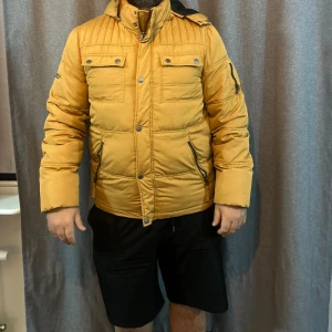 Gul pufferjacka från Hammond - Säljer en gul pufferjacka från Hammond med huva och stora tryckknappar framtill. Jackan har flera fickor med dragkedja och knappstängning, samt ett stort tryck med märket på ryggen. Perfekt för kalla dagar och har en schysst streetstil. Har en fläck som knappt syns 