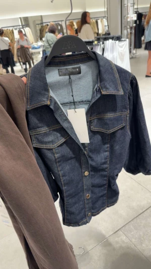 Mörkblå jeansskjorta från Zara - Snygg mörkblå jeansskjorta från Zara med klassisk krage, två bröstfickor med lock och kontrastsömmar. Skjortan har knappar framtill och lång ärm med lätt puffad axel. Perfekt för dig som gillar en cool och tidlös denimlook.