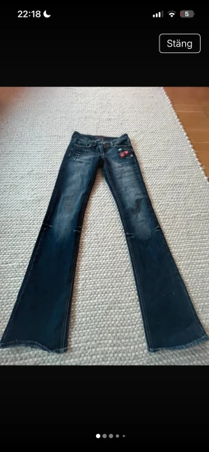 Mörkblå bootcut jeans med broderi - Snygga mörkblå jeans med bootcut passform och slitningar på benen. Jeansen har ett rött broderat märke på fickan och klassisk femficksdesign. Perfekta för dig som gillar en retro vibe och vill sticka ut lite extra. fråga gärna om fler bilder eller mått