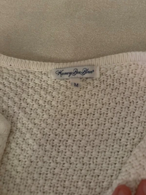 Vit stickad kofta från HoneyBeeBlue - Säljer en vit stickad kofta från HoneyBeeBlue i storlek M. Koftan har grovstickad struktur, rund halsringning och två snygga silverfärgade knappar framtill. Långa ärmar och praktiska fickor gör den både chill och stilren.