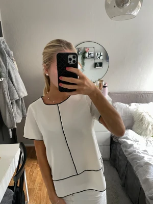 Vit blus med svarta kanter - Säljer en vit blus med kort ärm och svarta kontrastsömmar längs kanterna och i ett grafiskt mönster framtill. Blusen har en rak, lite loose passform och rund halsringning. Perfekt för en clean och stilren look.