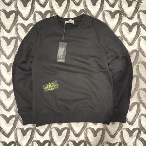 Svart sweatshirt från Stone Island - Svart sweatshirt från Stone Island med klassisk rund hals och ribbade muddar. Tröjan har det ikoniska Stone Island-märket på vänster ärm i gult och grönt. Materialet är mjuk bomull och passformen är normal. Perfekt för dig som gillar stilrena och exklusiva streetwear-plagg.