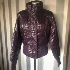 Lila pufferjacka från Terranova M - Snygg glansig pufferjacka från Terranova i mörklila. Jackan har hög krage, dragkedja och knappar framtill samt ribbade muddar i midja och ärmslut. Perfekt för dig som vill sticka ut med en cool och modern look.