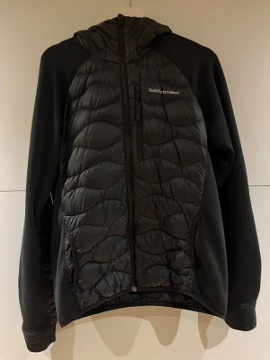 Svart pufferjacka Peak Performance - Svart pufferjacka från Peak Performance med huva och snygg quiltad framsida. Jackan har dragkedja hela vägen och två sidofickor. Materialet är en mix av polyester och syntet, perfekt för kyliga dagar. Loggan sitter på bröstet och designen är stilren och sportig.