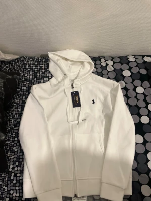 Vit hoodie från Polo Ralph Lauren - Vit hoodie från Polo Ralph Lauren med klassisk logga broderad på bröstet. Tröjan har huva med snörning, dragkedja framtill och två fickor. Mjuk bomullskvalitet och långärmad modell som passar perfekt för chill dagar.