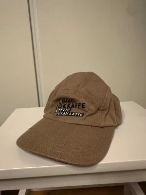 Lemmel keps  - Snygg brun keps från Lemmel med broderad text 'Lemmel Kokkaffe Ett liv utan latte' framtill. Kepsen har böjd skärm, justerbar rem med metallspänne. Perfekt för dig som gillar outdoor och vill sticka ut med en unik look. Nypris: 499kr