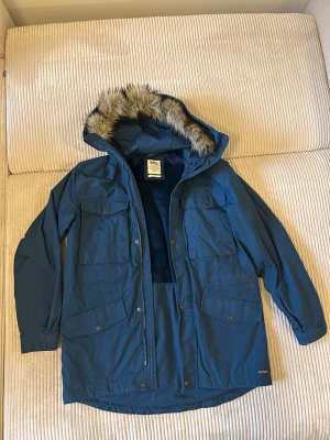 Blå parkajacka med päls från Fjällräven - Säljer en blå parkajacka från Fjällräven med fuskpäls på huvan och flera stora fickor framtill. Jackan är tillverkad i slitstark G-1000 polyester och har ett mjukt, mörkblått foder. Perfekt för kalla dagar och har både dragkedja och knappar framtill.