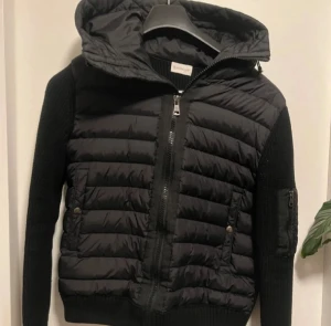 Svart Moncler pufferjacka med huva - Svart pufferjacka från Moncler med stickade ärmar och quiltad kropp. Jackan har en stor huva, dragkedja framtill och fickor med knappstängning. Moncler-logga på ärmen och en extra ficka med dragkedja på armen. Perfekt för dig som gillar stilren och sportig look.