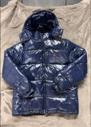 Marinblå glänsig pufferjacka från Polo Ralph Lauren - Säljer en marinblå pufferjacka från Polo Ralph Lauren med blank finish och röd broderad logga på bröstet. Jackan har huva, dragkedja framtill och elastiska muddar. Perfekt för kalla dagar och riktigt snygg streetstil. Storlek M men sitter lite stor i storleken! Pris går att diskutera! 💯🫡
