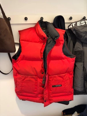 Röd dunväst från Canada Goose - Säljer en röd dunväst från Canada Goose med svart foder och hög krage. Västen har två stora fickor framtill, dragkedja med logotypdetalj och tryckknappar. Perfekt för kalla dagar och har en klassisk, puffad look som sticker ut. Aldrig använd så hör av er vid minsta funderingar. ‼️priset är inte hugget i sten men rimligt pris för nyskick kostar den 8000‼️STL M men passar S