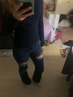 Mörkblå bootcut jeans med slitningar - Snygga mörkblå jeans med hål på låren och knäna. Bootcut-modell med vita sömmar och klassiska fickor både fram och bak. Jeansen har låg midja och är tillverkade i jeansmaterial. Från ginatricot