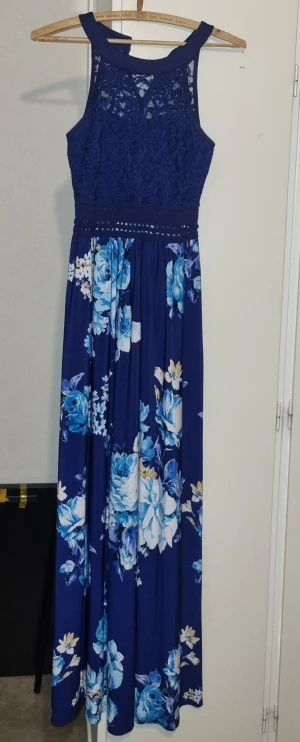 Marinblå långklänning med spets och blommor - Snygg marinblå långklänning med spets upptill och vackert blommigt mönster i blått och vitt på kjolen. Klänningen har ärmlös design och markerad midja med dekorativt band. Perfekt för dig som vill sticka ut med en elegant och romantisk look.