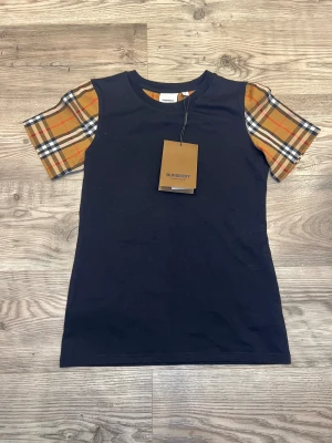 Burberry t-shirt med rutiga ärmar - Svart t-shirt från Burberry med klassiska rutiga korta ärmar i beige, rött och svart. T-shirten har rund halsringning och är gjord i mjuk bomull. Perfekt för dig som vill ha en stilren look med en touch av lyxig streetstyle.