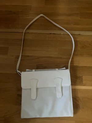 Vit axelväska i skinnimitation - Stilren vit axelväska med magnetlås och justerbar rem. Väskan har ett stort huvudfack, innerficka med dragkedja och en ytterficka med dragkedja på baksidan. Snyggt lock med dekorativa detaljer framtill. Perfekt för dig som gillar minimalistisk och clean stil.