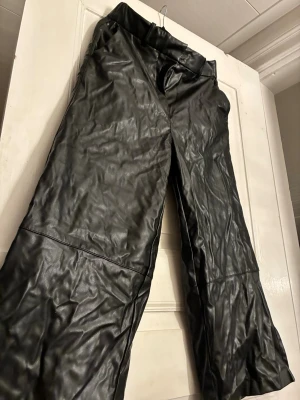Svarta vida byxor i fejkskinn från H&M - Säljer ett par svarta byxor i fejkskinn från H&M, storlek 44. Byxorna har bred resår i midjan, vida ben och raka sömmar över knäna. Snyggt glansigt material som ger en edgy look. Perfekta till sneakers eller boots.