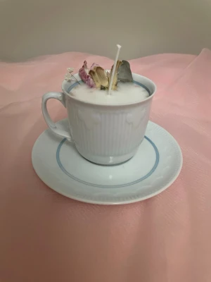 Doftljus i vaniljdoft i en vit tekopp med blå rosett och blommor - Det här ljuset är gjort av återanvändbara material. Tekoppen är köpt secondhand och stearinet är från kyrkor som tänkte slänga det. Det här ljuset har en svag doft utav vanilj, så känsliga även kan använda det. Det här ljuset har även torkade blommor på som dekoration.